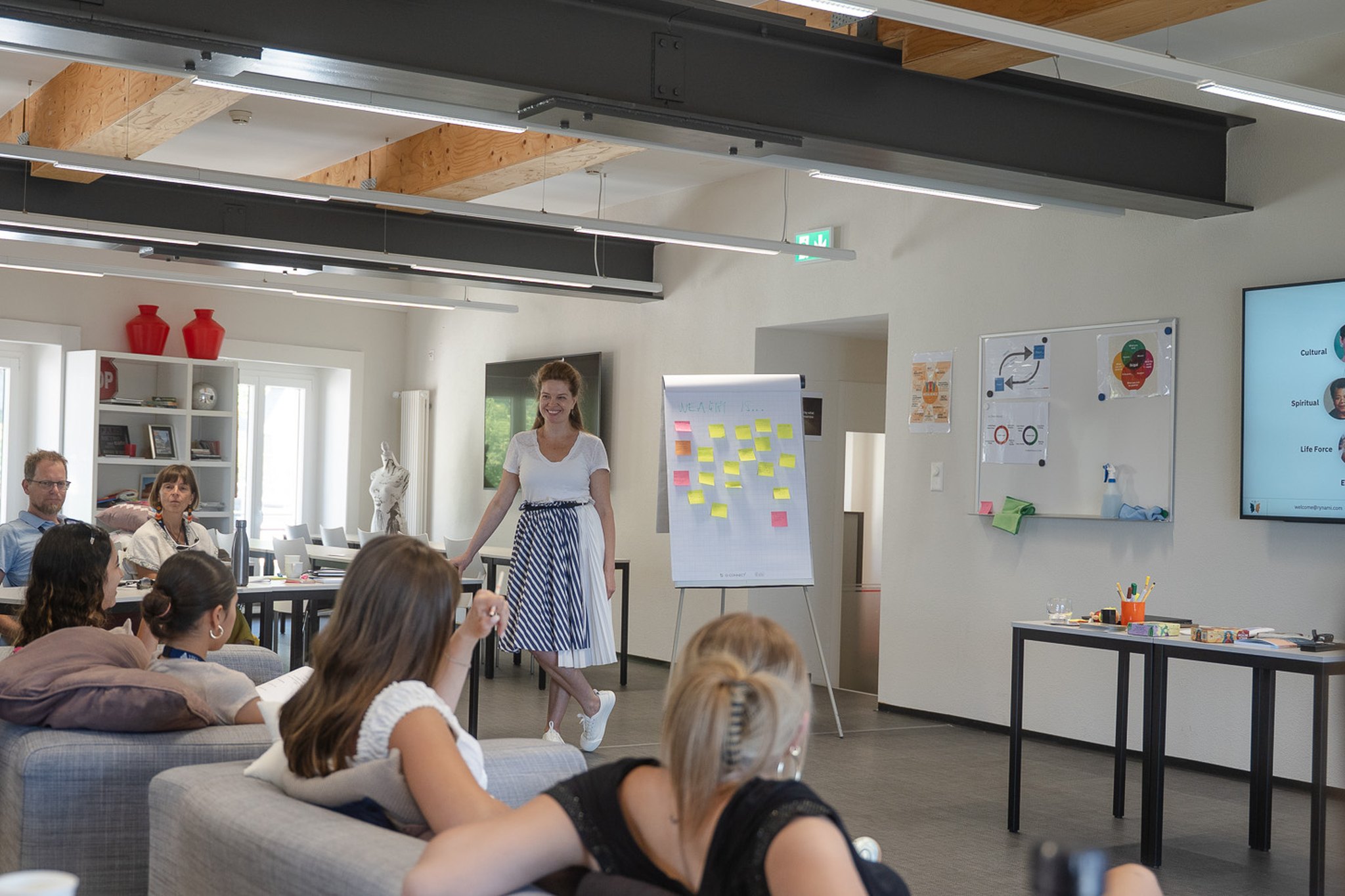 Ryna Mi at the flip chart, a group of participants on sofas and at tables, a session of The Wealth School
